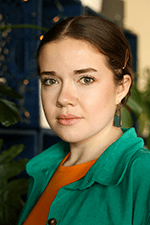 A portrait of a person in their mid-20s. Their brown hair is pulled back into a bun. They wear an orange shirt under an aquamarine jacket.