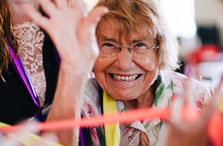 An older woman with a sandy blond hair and wire frame glasses in a flower print shirt smiles and waves at the camera. Colorful ribbons are streamed between her and two others who stand on either side of her.